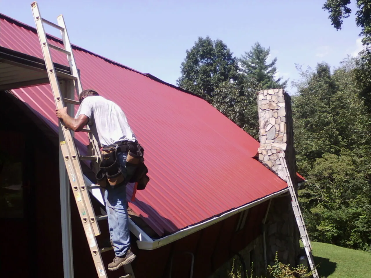 Roofer on a ladder beside a red metal roof and stone chimney — Soffit & Fascia Repair by Roofing Brothers in Leavittsburg